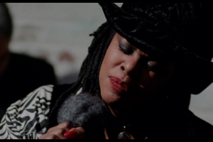 Still from Q Lazzarus performing in the movie “Philadelphia”. Q, an African-American woman, is wearing a black hat and holding a microphone she is singing into.