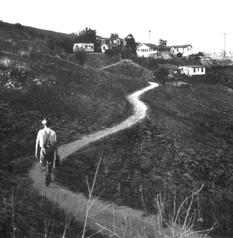 Black and white image of a man, back to us, walking up a dirt path towards a house