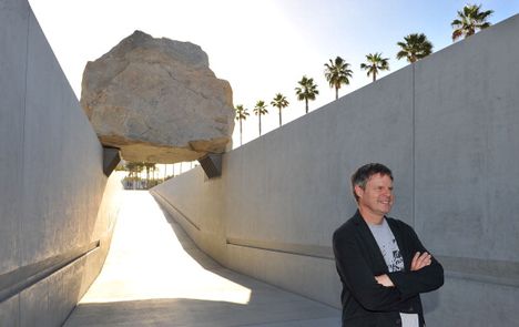 A middle-aged white man stands with his arms crossed with an art installation behind him