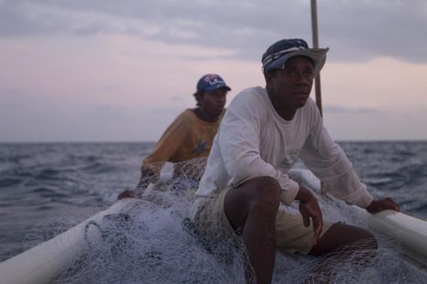 Two young black men sit in a boat with nets around them, looking into the distance