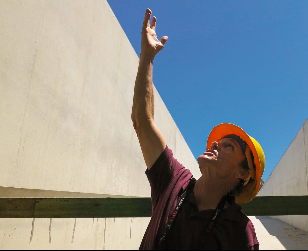 Artist Michael Heizer, a middle-aged white man in a maroon shirt and orange hard hat, looks and indicates upwards