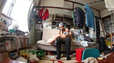 A man with a toddler on his lap sits in a living room filled with clothes and toys 