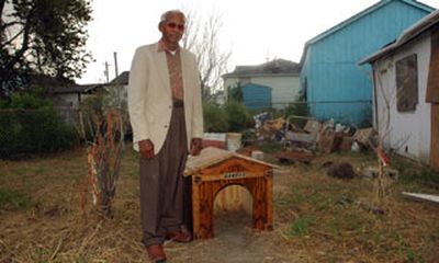 An older black man in slacks and a jacket stands in a unkept yard  