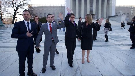 Two men holding hands stand next to two women holding hands, all in suits, in front of a government building