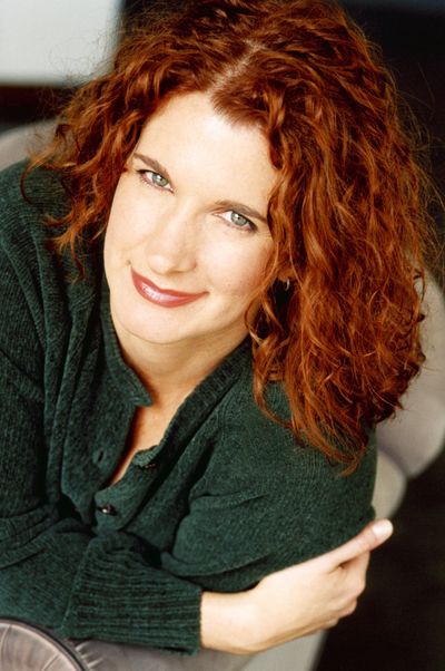A white woman with curly red hair wears a green sweater and looks up at the camera, smiling