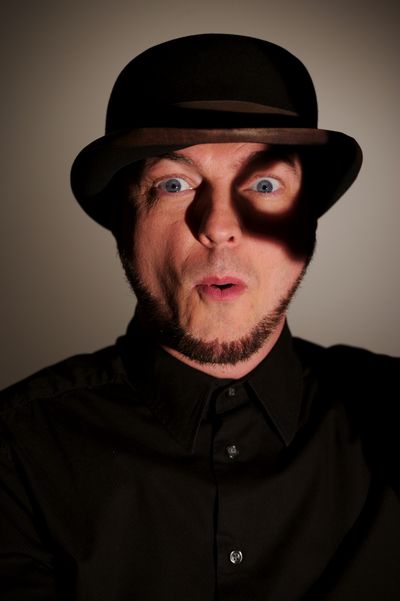 A white man in a bowler hat and a black button down shirt in front of a gray background, with a shadow around their eye