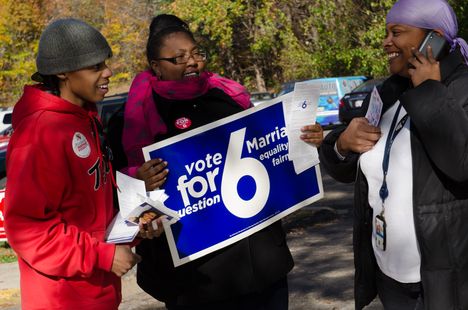 Two young black women and one young black man standing outside, all smiling, with woman on a phone call and another woman holding a campaign sign that reads "Vote For Question 6"