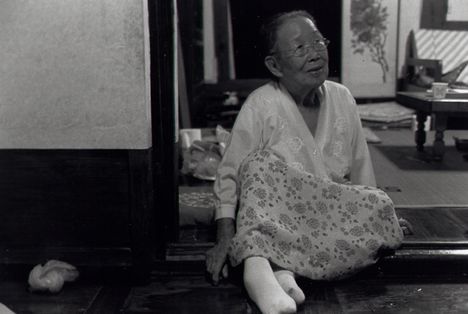 Black and white image of an older Asian woman sitting on the floor of a home