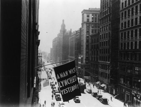 Black and white image from a window of a city street with a flag hanging off a building reading "A Man Was Lynched Yesterday"