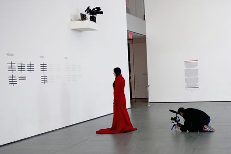 A woman in a long red dress stars at a piece of artwork as a man with a camera kneels behind her, filming