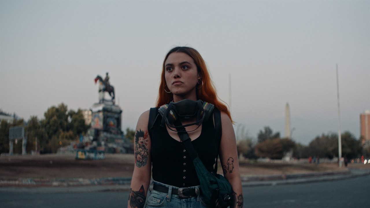 A tattooed Chiliean woman, wearing a black tank top and carrying a shoulder bag, stands in the middle of a city street, with monuments in the background. From Franz Böhm’s 'Dear Future Children', courtesy of Hot Docs.