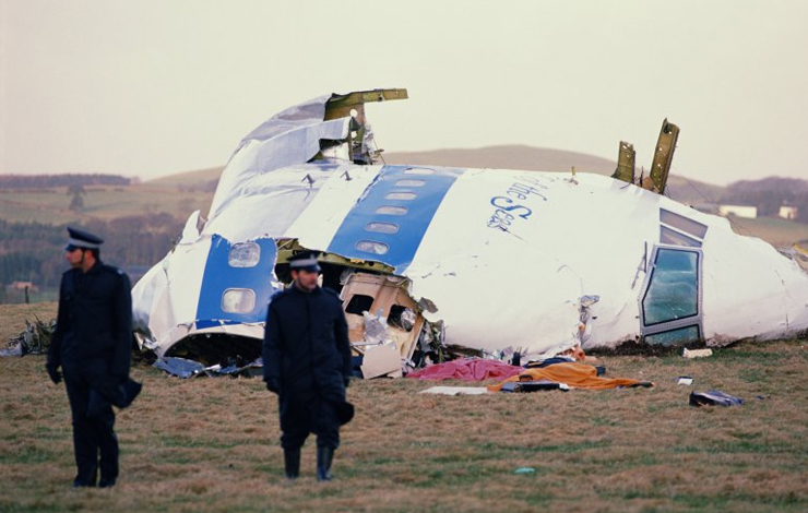 Two men in black coats and hats stand before a crashed plane. 
