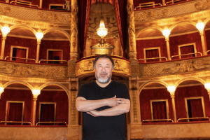 An Asian man wearing a black shirt stands with his arms crossed in front of the balcony seating of an opera house