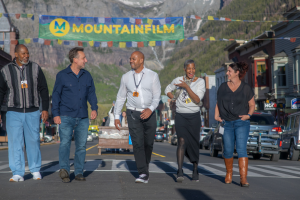 Image of five people walking together smiling and laughing down a street with a green and yellow banner behind them with text MOUNTAINFILM. Buildings line the side of the road and a big mountain is in the background. Image courtesy of Julian Rubinstein.