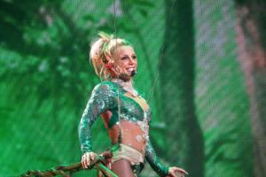 Pop star Britney Spears, subject the recnt documentary 'Framing Britney Spears," is pictured performing on stage, wearing an emerald green sequin outfit, singing into a wireless headset, clutching a brown railing, against a mesh, emerald green patterned backdrop.