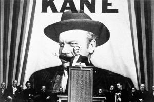 A man stands on a podium with a giant poster that reads 'Kane' in the back. Most first-time viewers of 'Citizen Kane'  don't realize that the News on the March short that opens the film is a clever parody of a series of theatrical shorts produced by Time, Inc. 