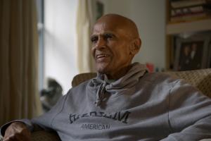 Harry Belafonte is a Black man, bald, smiling, wearing a grey hoodie and sitting on a chair, in front of a bookshelf. From Elvis Mitchell’s ‘Is That Black Enough For You?!?’ Courtesy of netflix