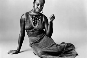 Nina Simone is sitting on the floor with her right arm propping her up. She is wearing a maxi knit dress, large decorative necklace.
