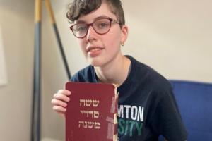 An image of non-binary teen AJ Hack holding his family's copy of the Mishnah, a red book with Hebrew lettering and a taped-together binding. 