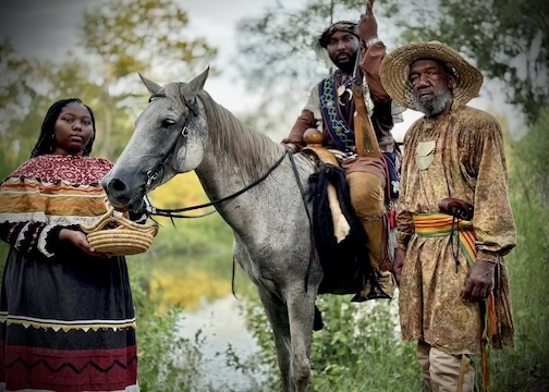 Three Black Seminoles, a woman and two men, wearing traditional clothing. One of the men is sitting on a gray horse while the others stand next to the horse.