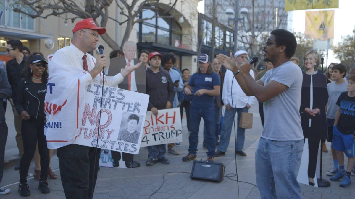 A Trump supporter wrapped in a MAGA flag debates with an opponent in front of a crowd on the street