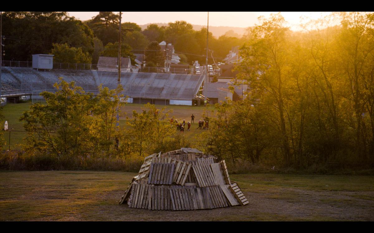 a pile of woodpallets sits on a hill above a pop-warner football practice at sunset.
