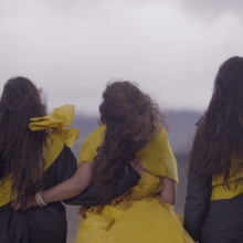 Five women stand with their backs to the camera embracing each other in a line with the gray sky in front of them. They all have long brown hair and are wearing traditional Native Hawaiian kihei dyed with olena (turmeric) that are tied around their shoulders. 