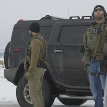 Two FBI agents in gear standing next to a big black armored SUV