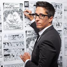 Alison Bechdel is a white woman with short hair black-rimmed glasses. She is wearing a black suit and is holding a marker to a wall of her comics. Image from Vivian Kleinman's ‘No Straight Lines: The Rise of Queer Comics’. Courtesy of Outfest.