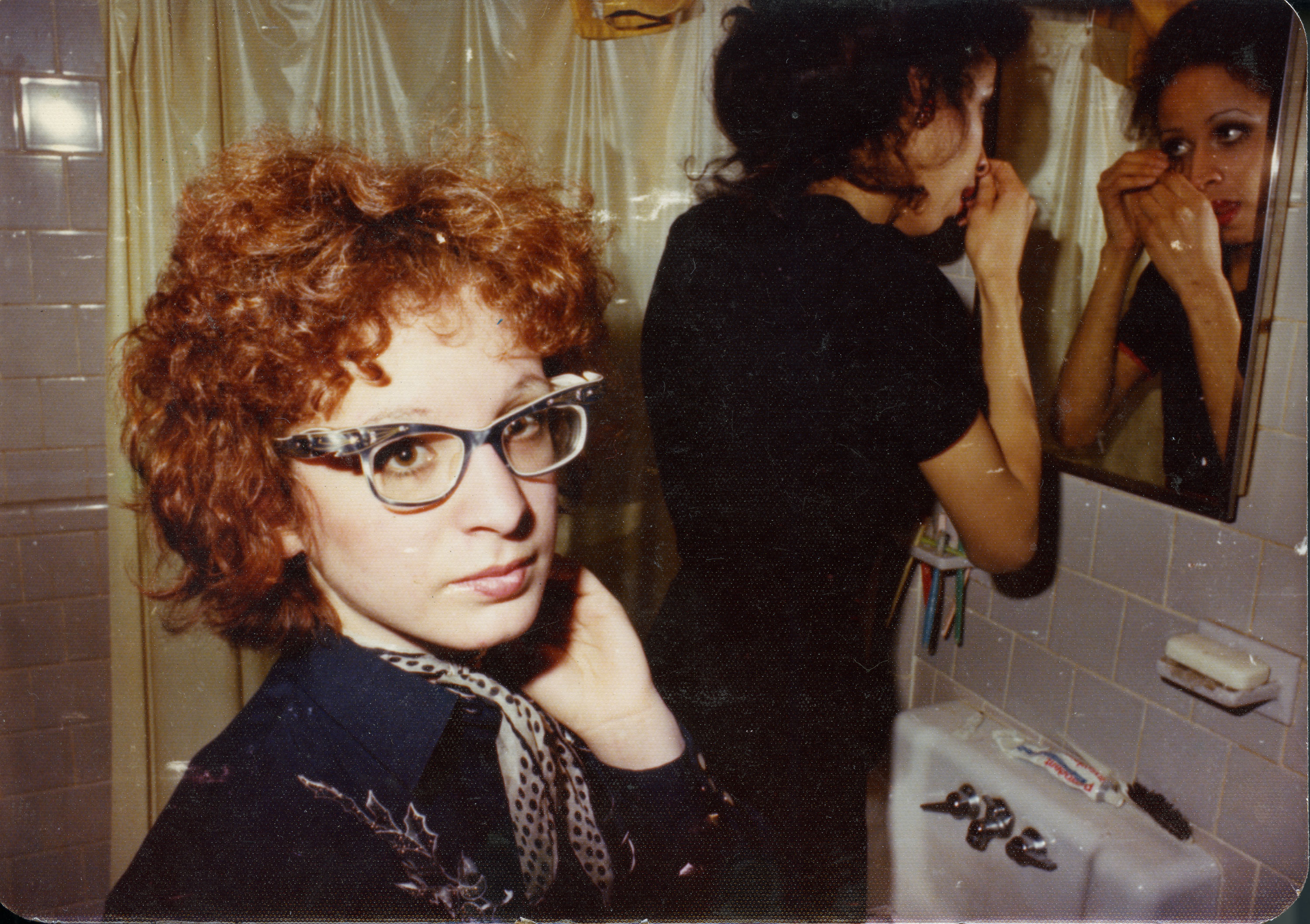 A woman wearing black adjusts her eye makeup in the mirror, while a red-headed woman wearing glasses looks at the camera in the foreground. From Laura Poitras' ‘All the Beauty and the Bloodshed.’ Photo courtesy of Neon.