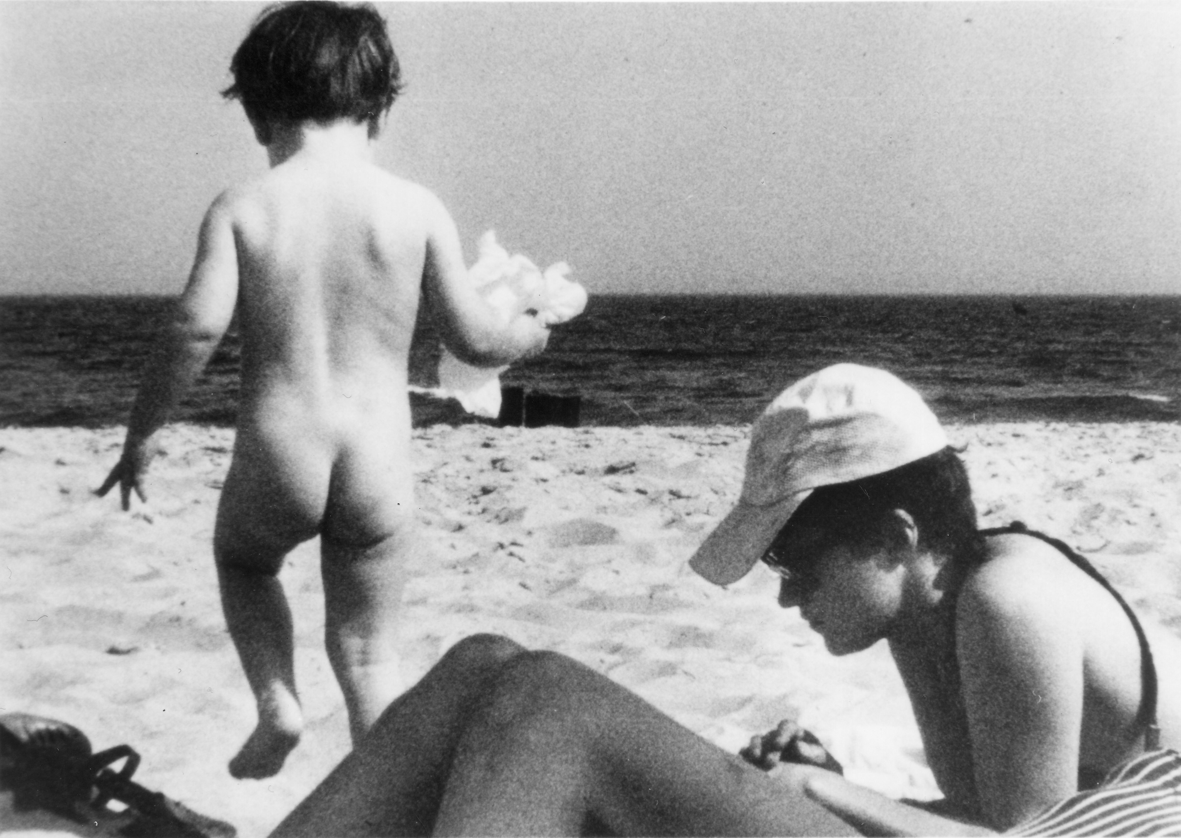 Black and white photo of a naked baby running on the beach. Adult person is laying in the sand on stomach wearing a baseball cap, swimsuit, and sunglasses.