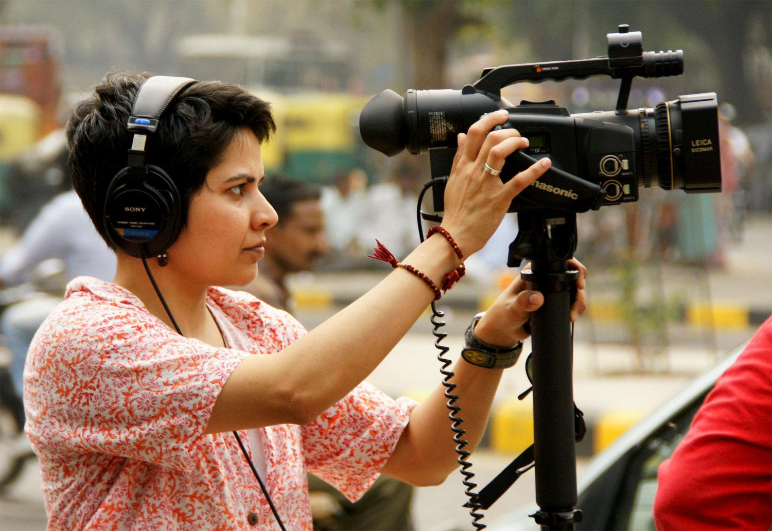 A Brown, badass, butch dyke is shooting a scene with queer rights protesters (offscreen) on the streets of New Delhi, with a video camera on a monopod and headphones covering her ears.