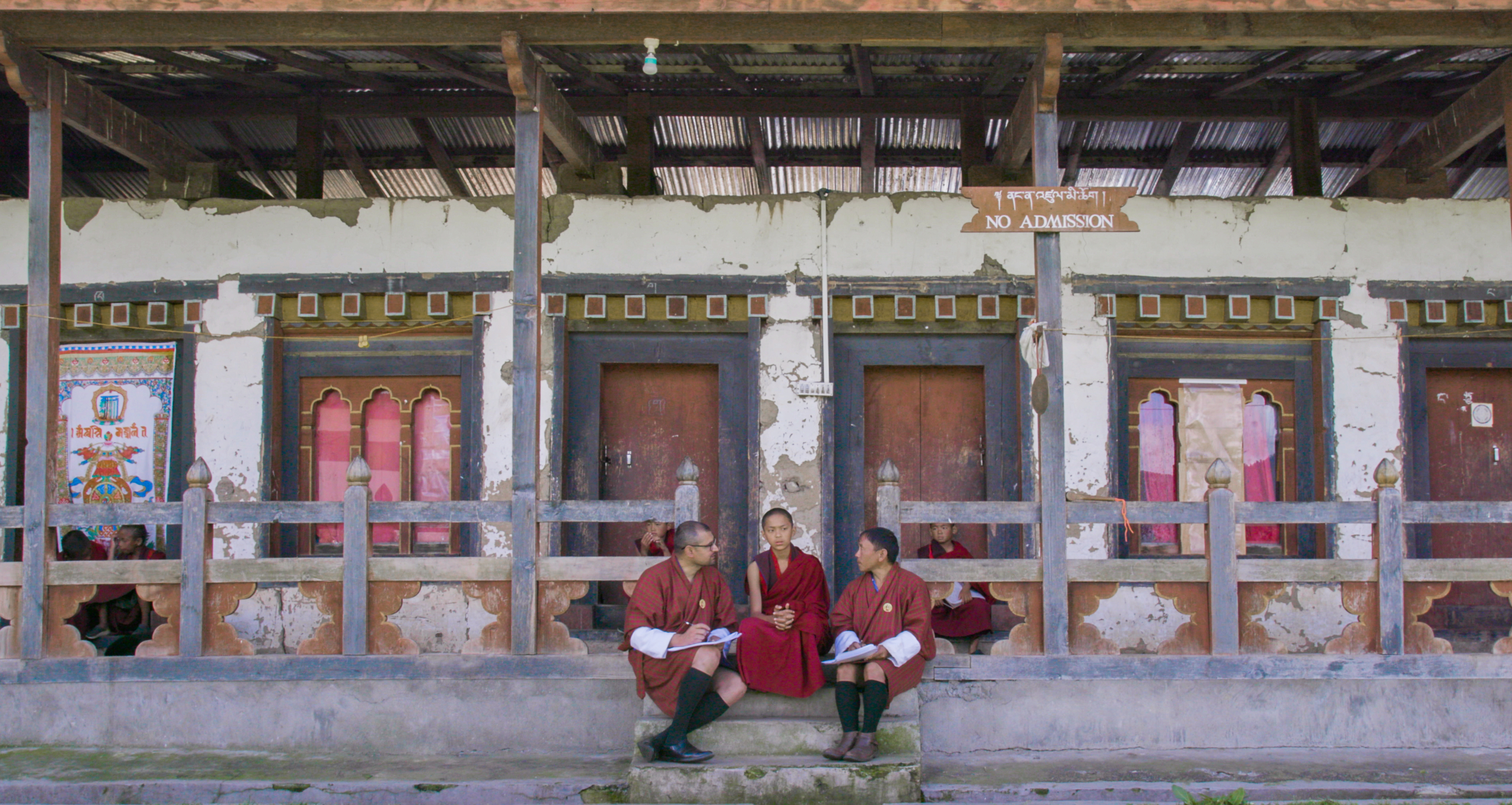 Still image from 'Agent of Happiness,' depicting three male figures seated on the steps of an ornate temple. All three are wearing robes in a deep maroon.