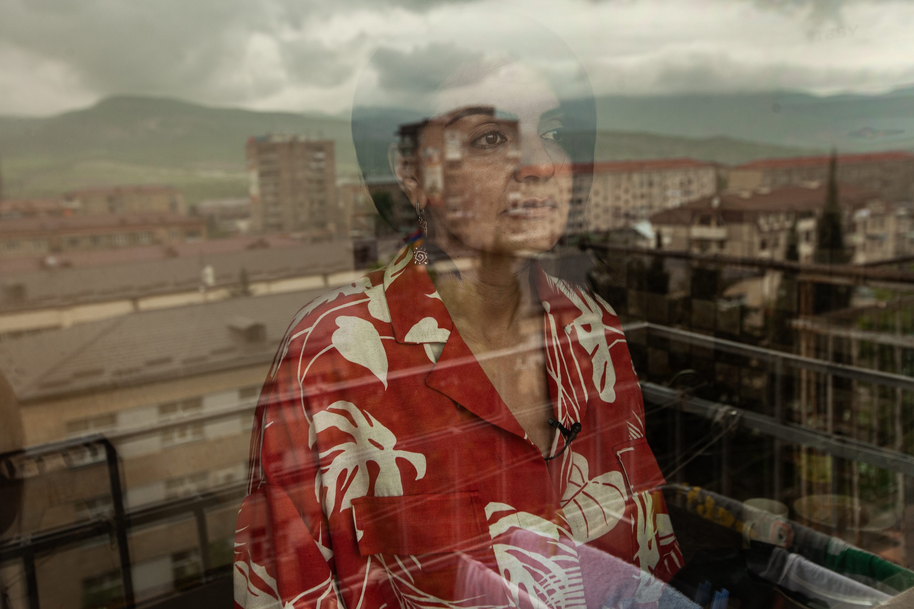 A woman wearing a patterned red shirt stands in a window, which shows a reflection of a rugged mountainous landscape.