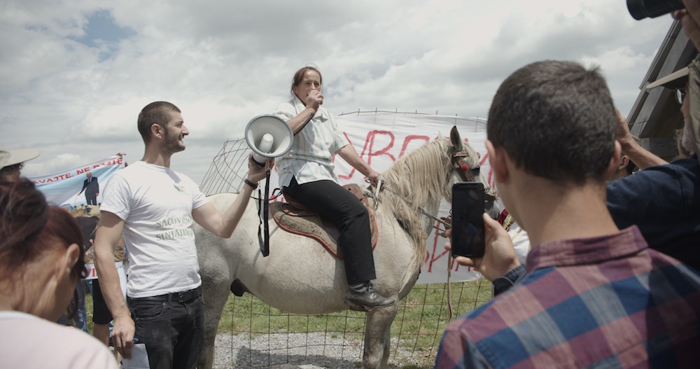 Person on a horse talking to a crowd with a banner in the background. With a person handing them a megaphone