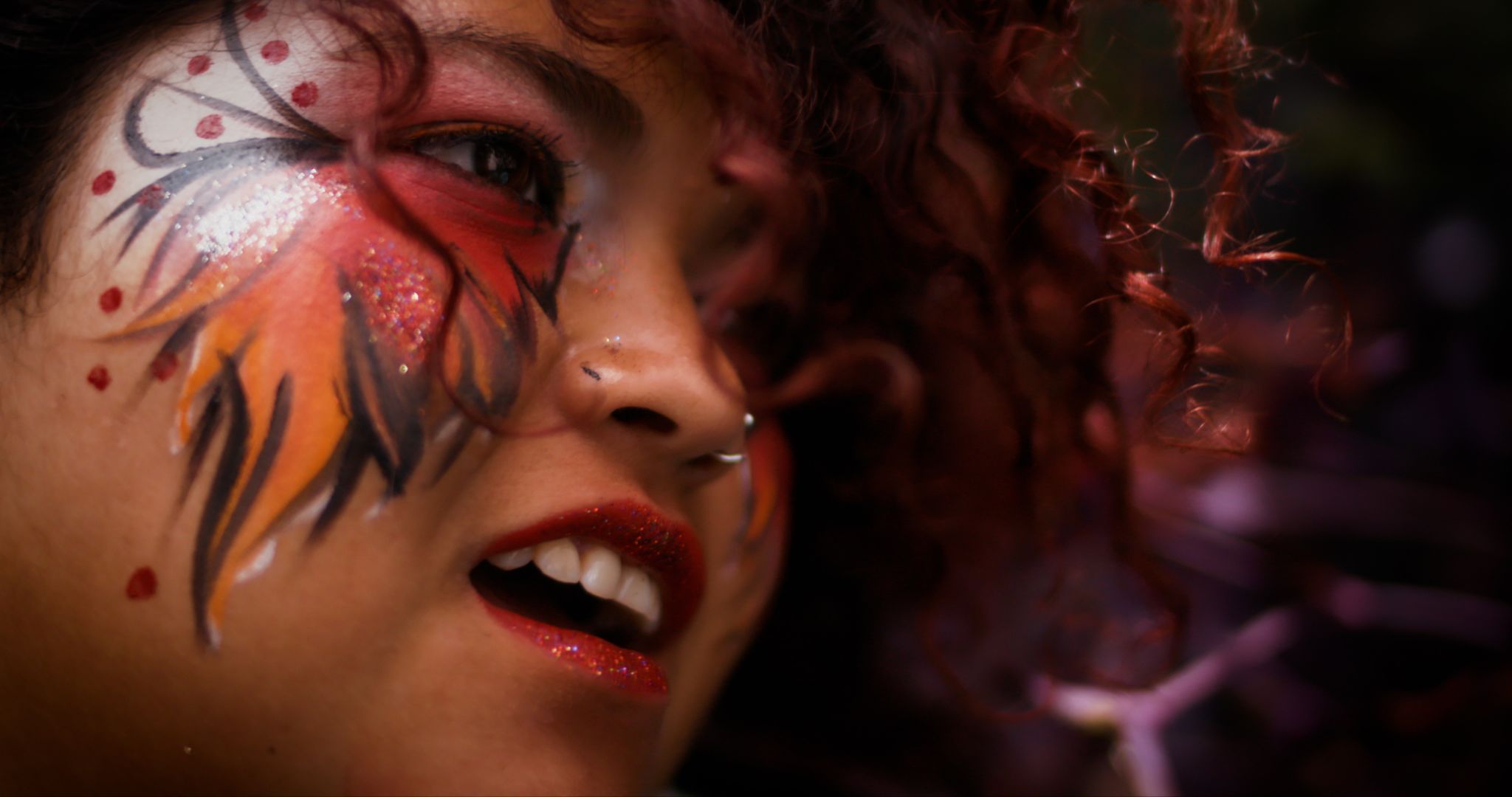 A close-up of a girl in bright glittery orange, red, and black facepaint. The focus is on her face, and the background is a vibrant purple. 
