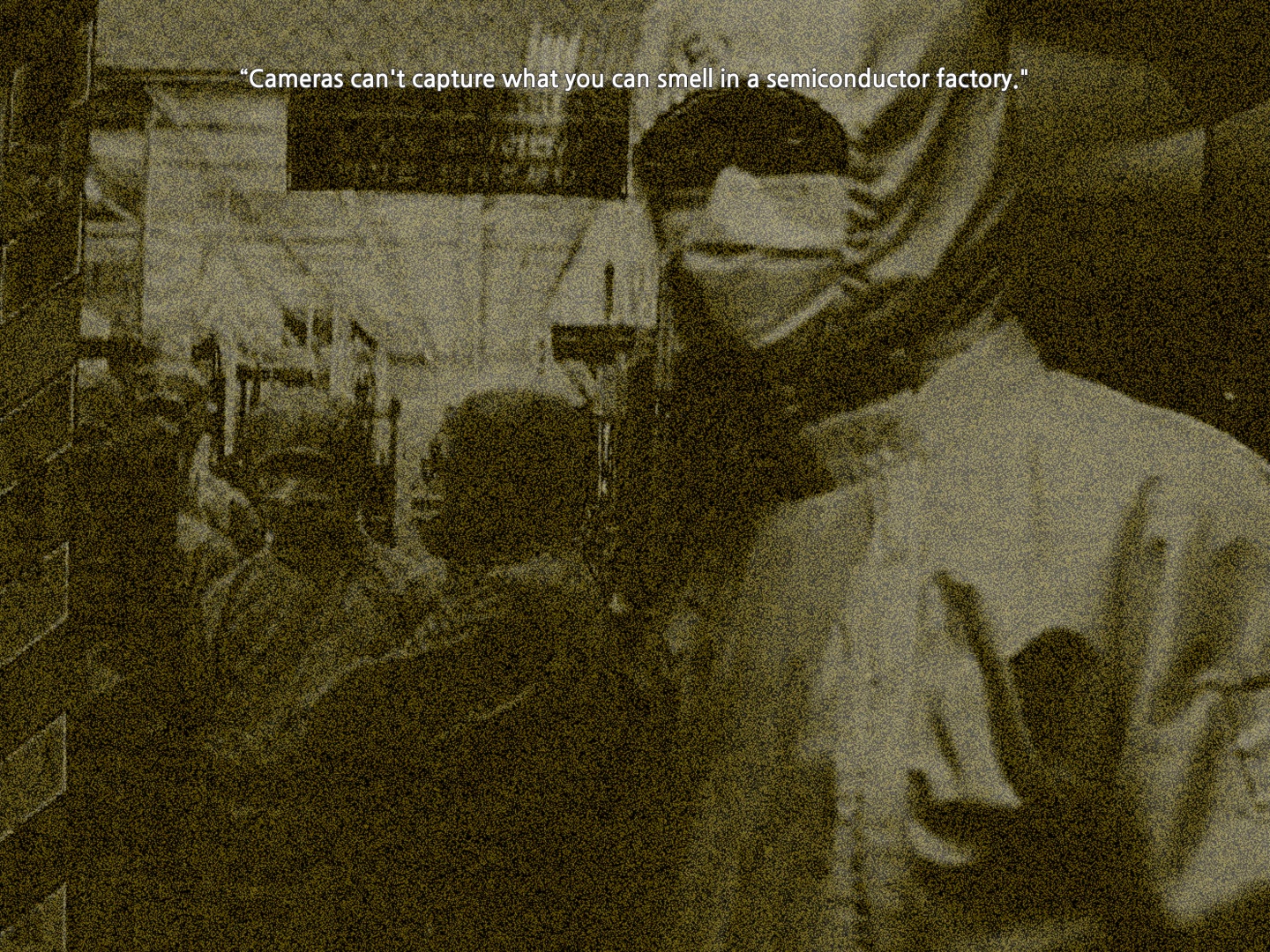 A person in safety gear and a mask looks at the camera, standing next to the door to a room where more people in the same gear are pictured inside.