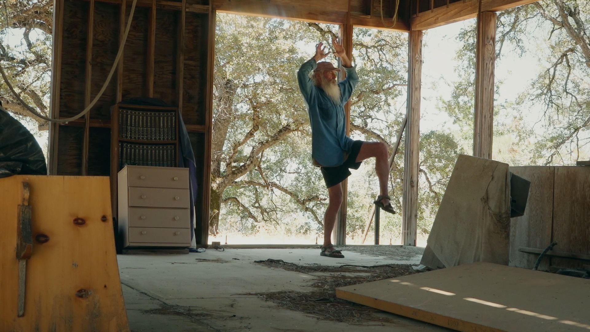 In an abandoned-looking shell of a house, a man with a long white beard does some stretches.