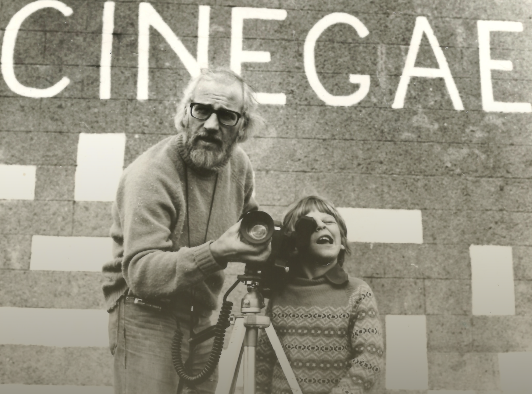 In front of a wall with "CINEGAEL" painted on it stands a father and his son, with a camera on a tripod.