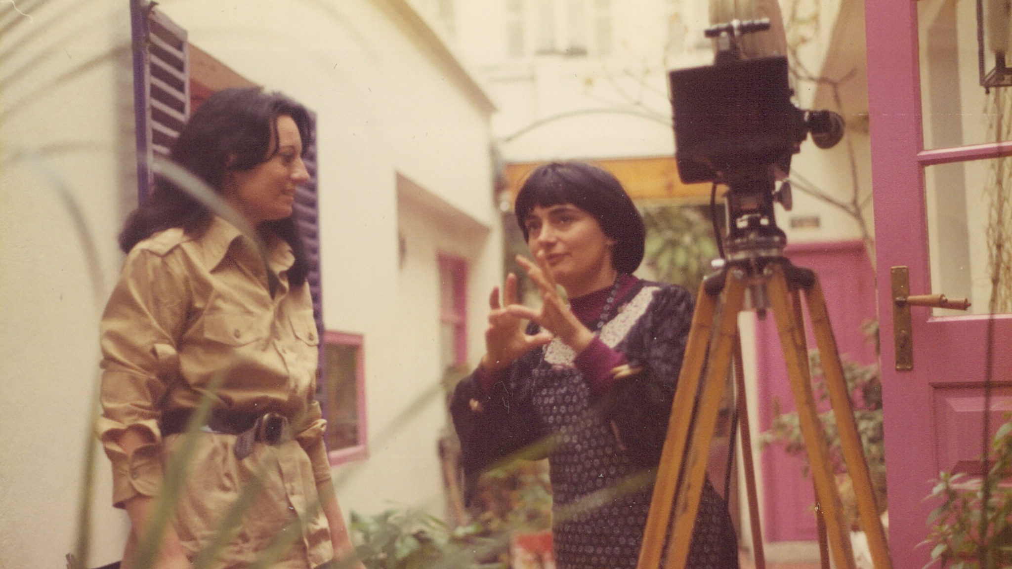A candid shot of a woman in a khaki outfit with long brunette hair(Raganelli) standing next to a woman with a bowl cut(Agnes Varda) as they converse. 