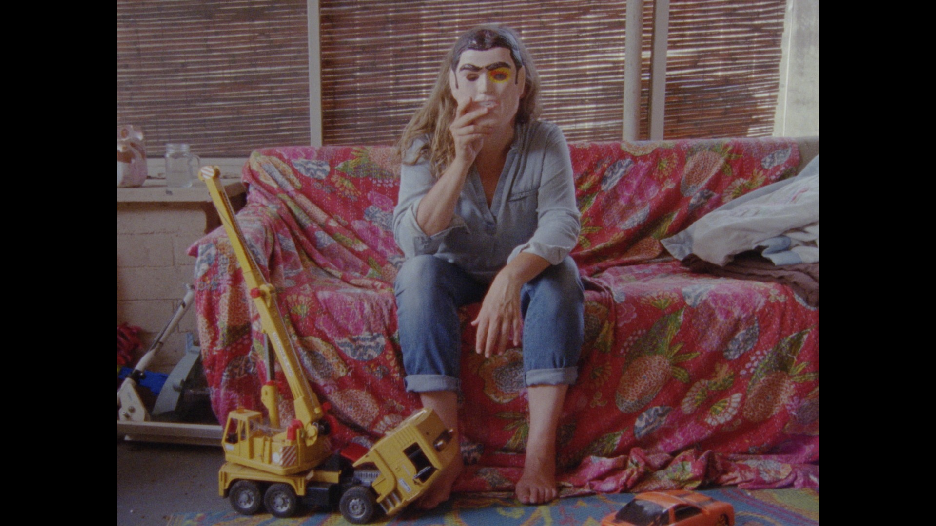 On a cloth-covered couch on an outdoor porch, a woman holds a Halloween mask over her face.