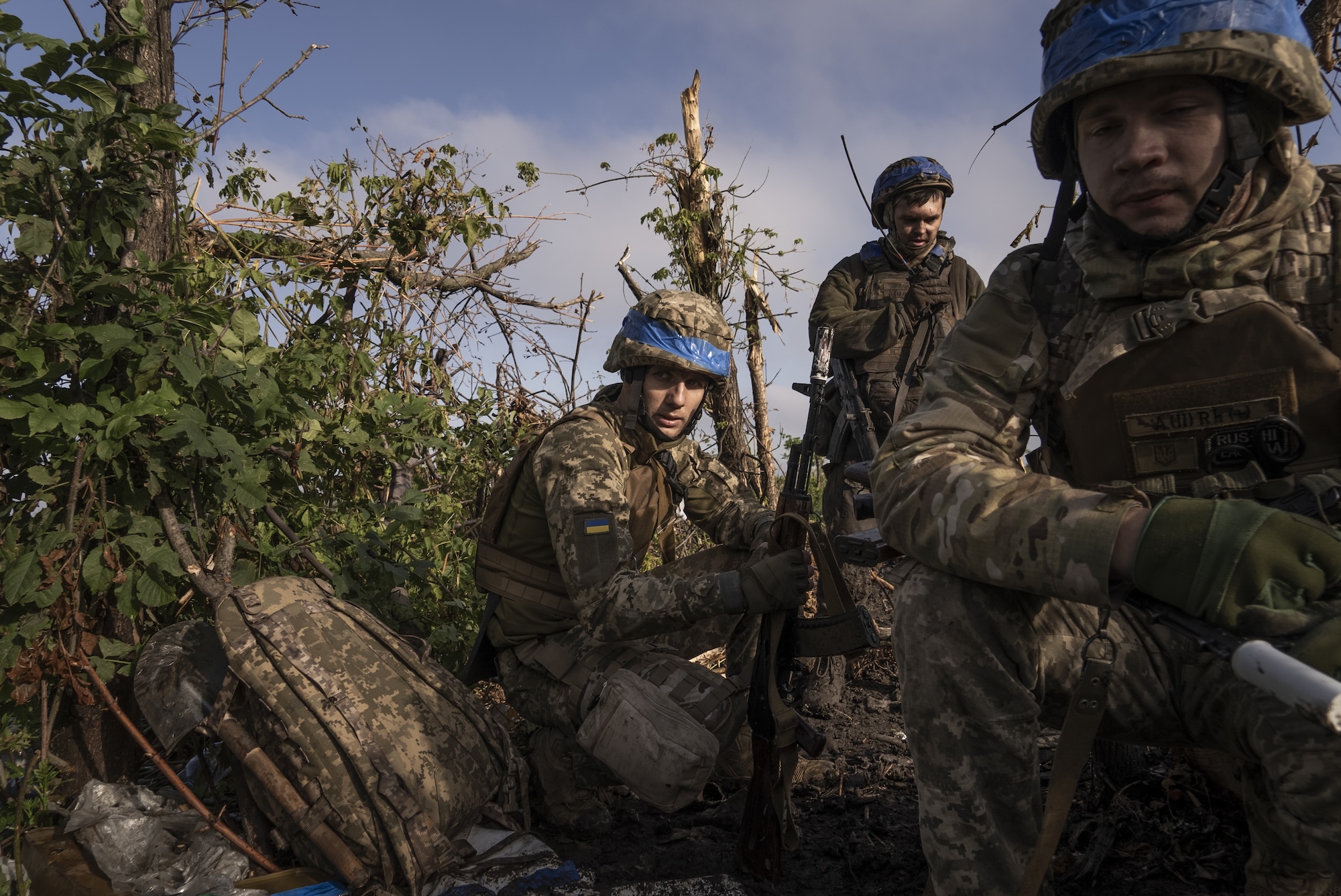 Three soldiers, with helmets and a small Ukrainian flag on their uniforms, crouching holding their guns outdoors