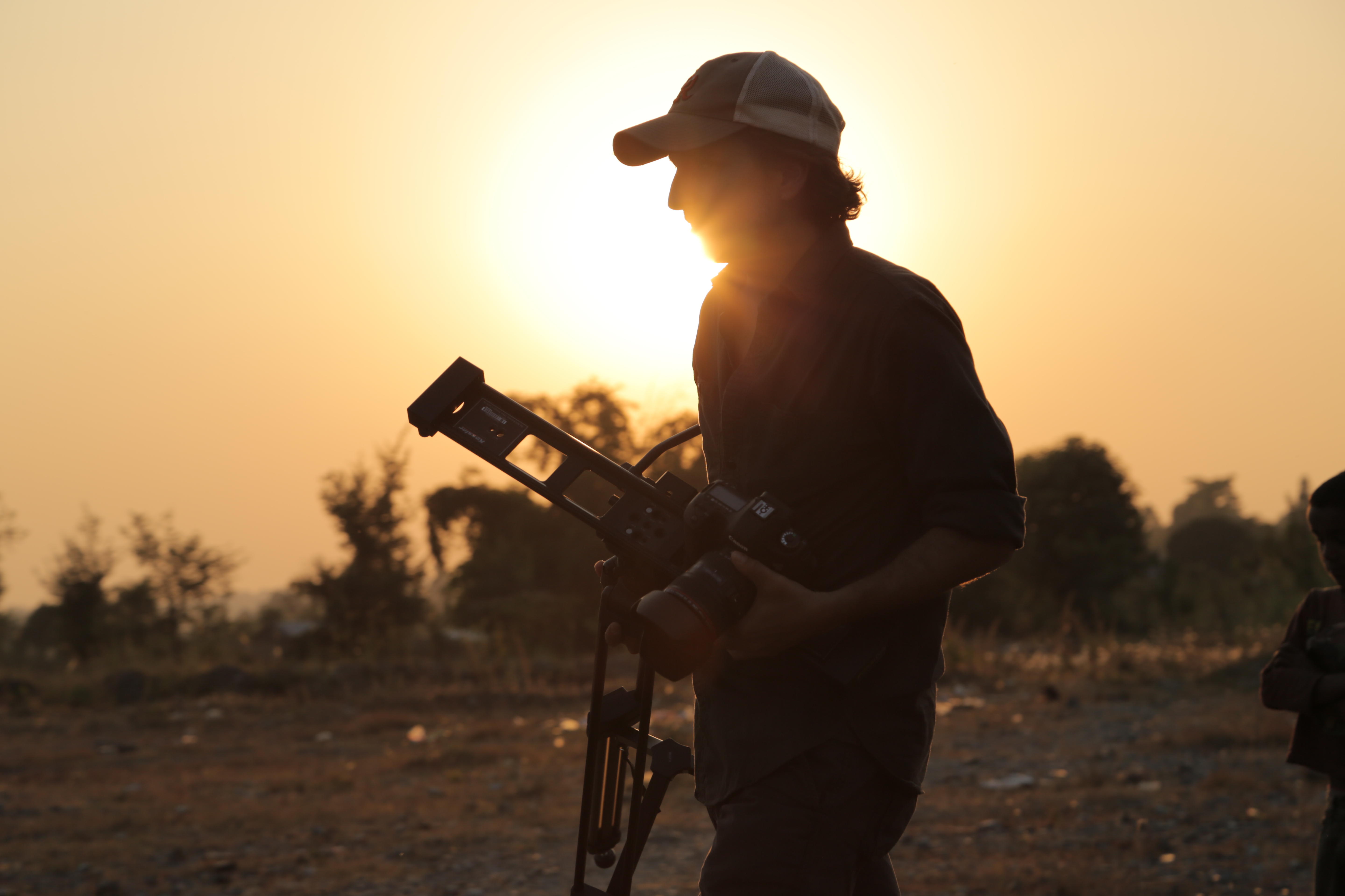 A man wearing a baseball cap and carrying a camera and tripod strolls through an arid landscape, illuminated by a sunset.