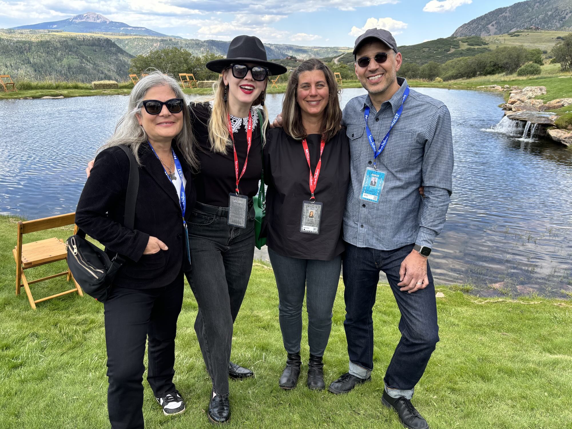 On the grassy banks of a lake, four white people wearing festival lanyards stand casually and happily.