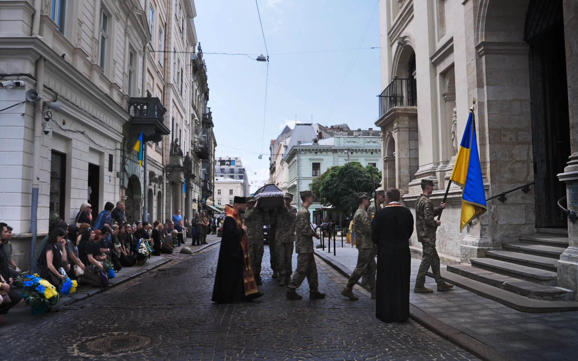 Led by a soldier carrying the Ukrainian flag, a casket is being carried by soldiers into a church; some onlookers kneel on the street as if it were a parade.