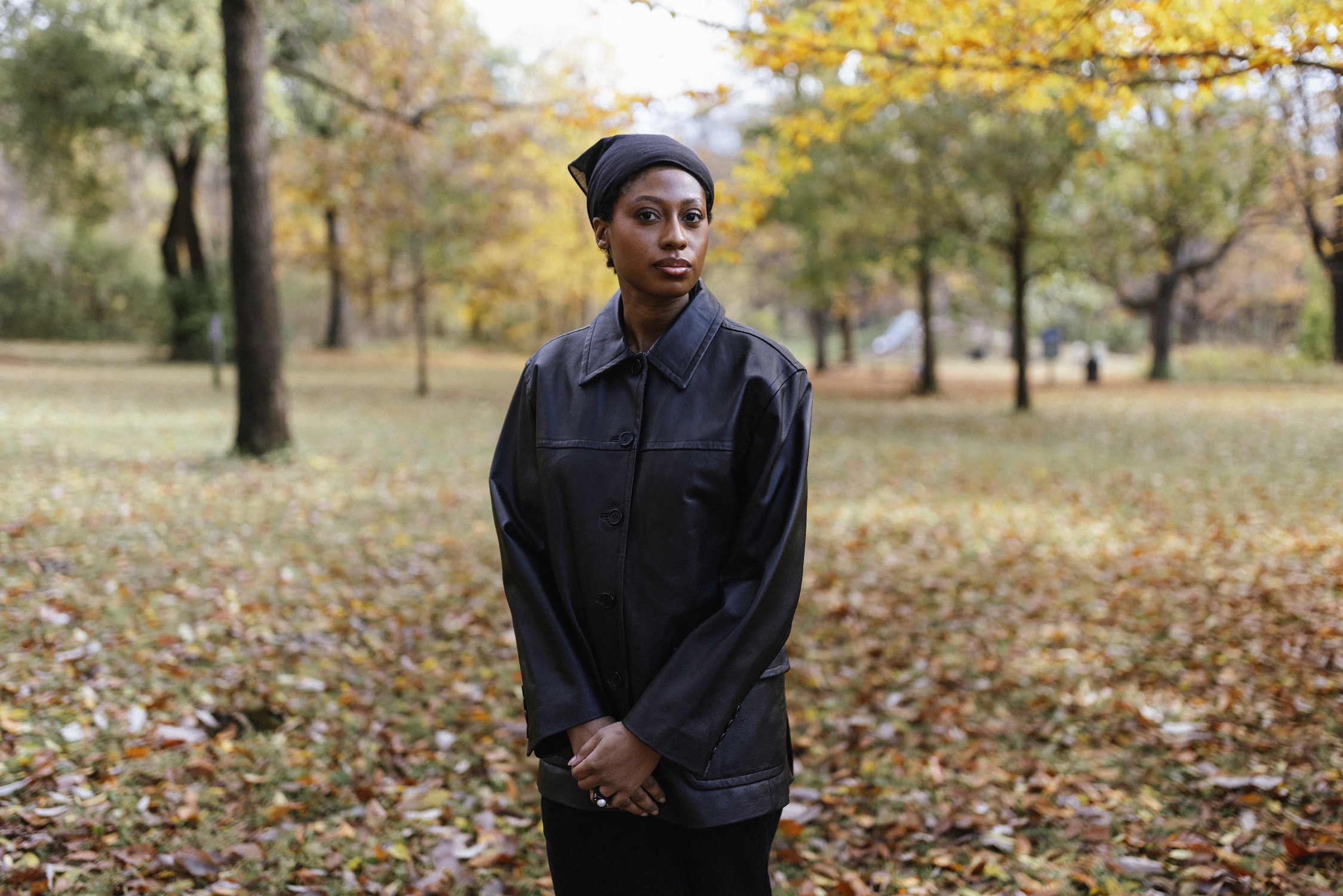 A Black woman in a black leather jacket stands with hands clasped in a field of autumnal leaves.