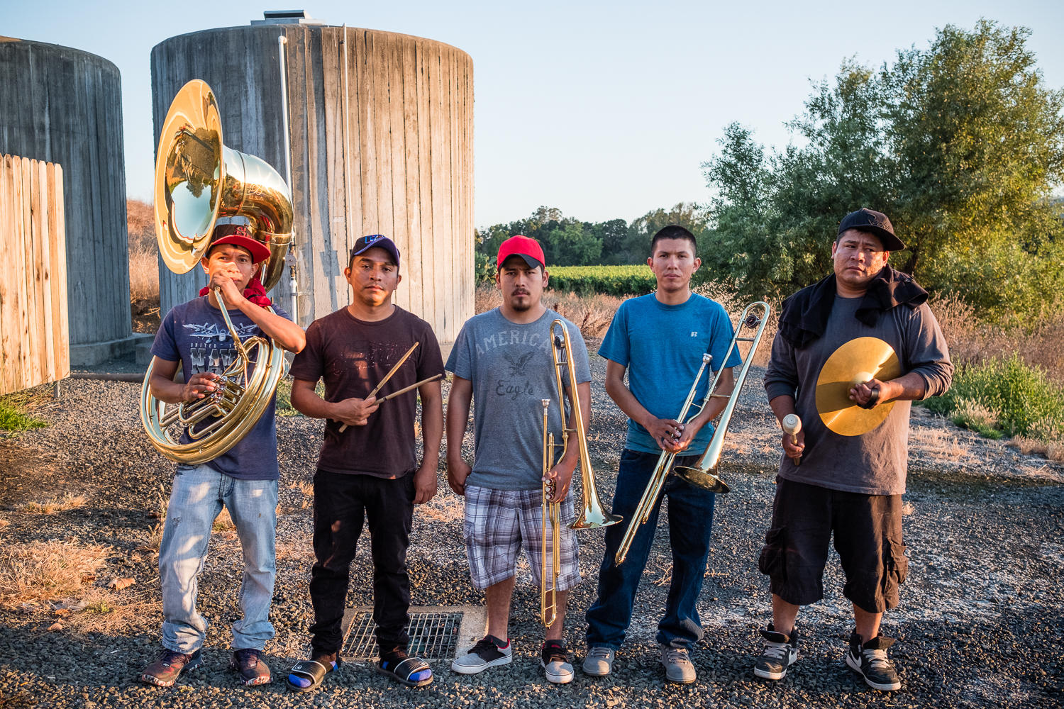 Five young men, each holding a different musical instrument, stand outside in a sunny field