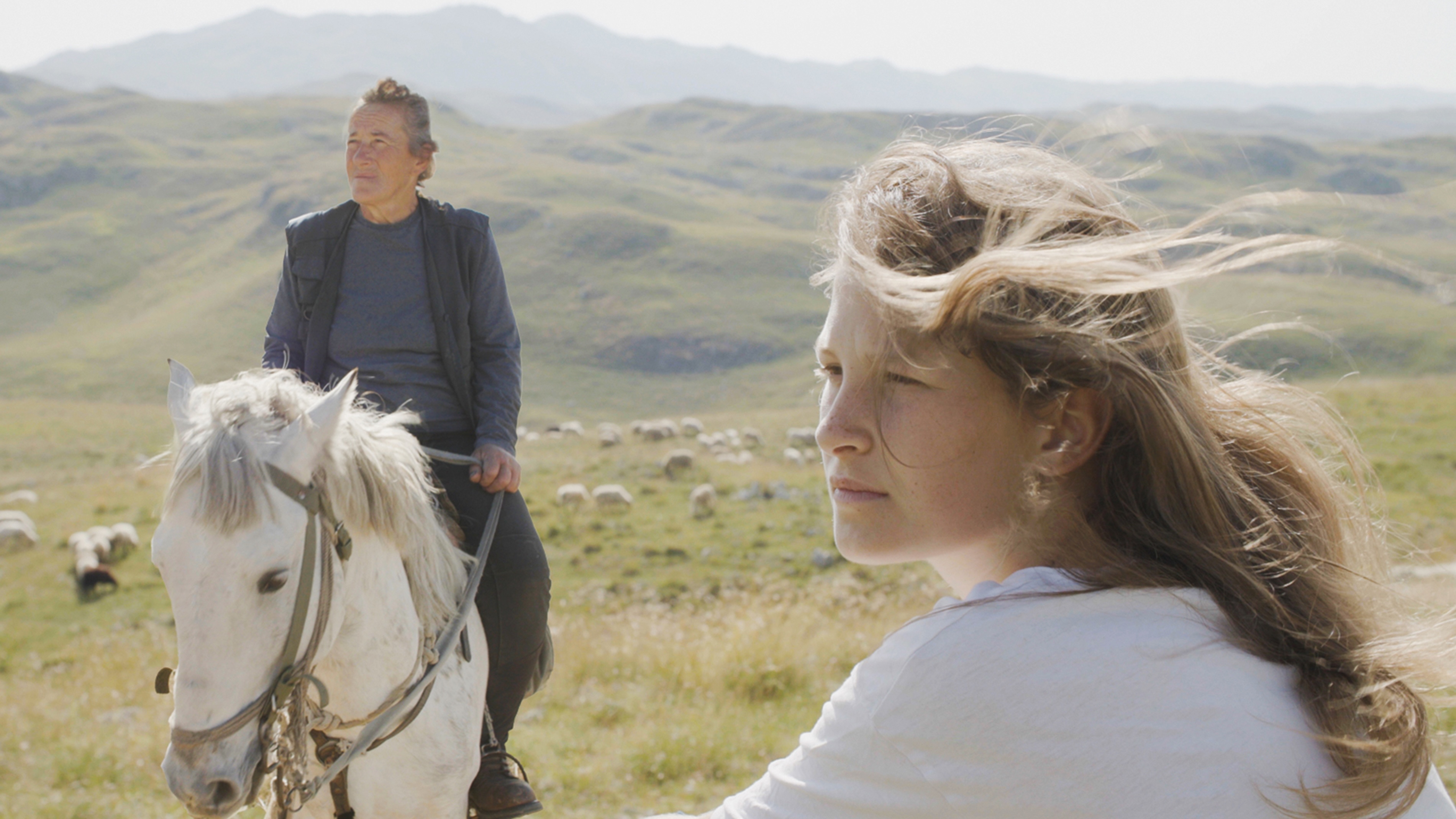 A young white blond girl in the mountains is seen in profile, while an older white woman with her grey hair pulled back rides a white horse