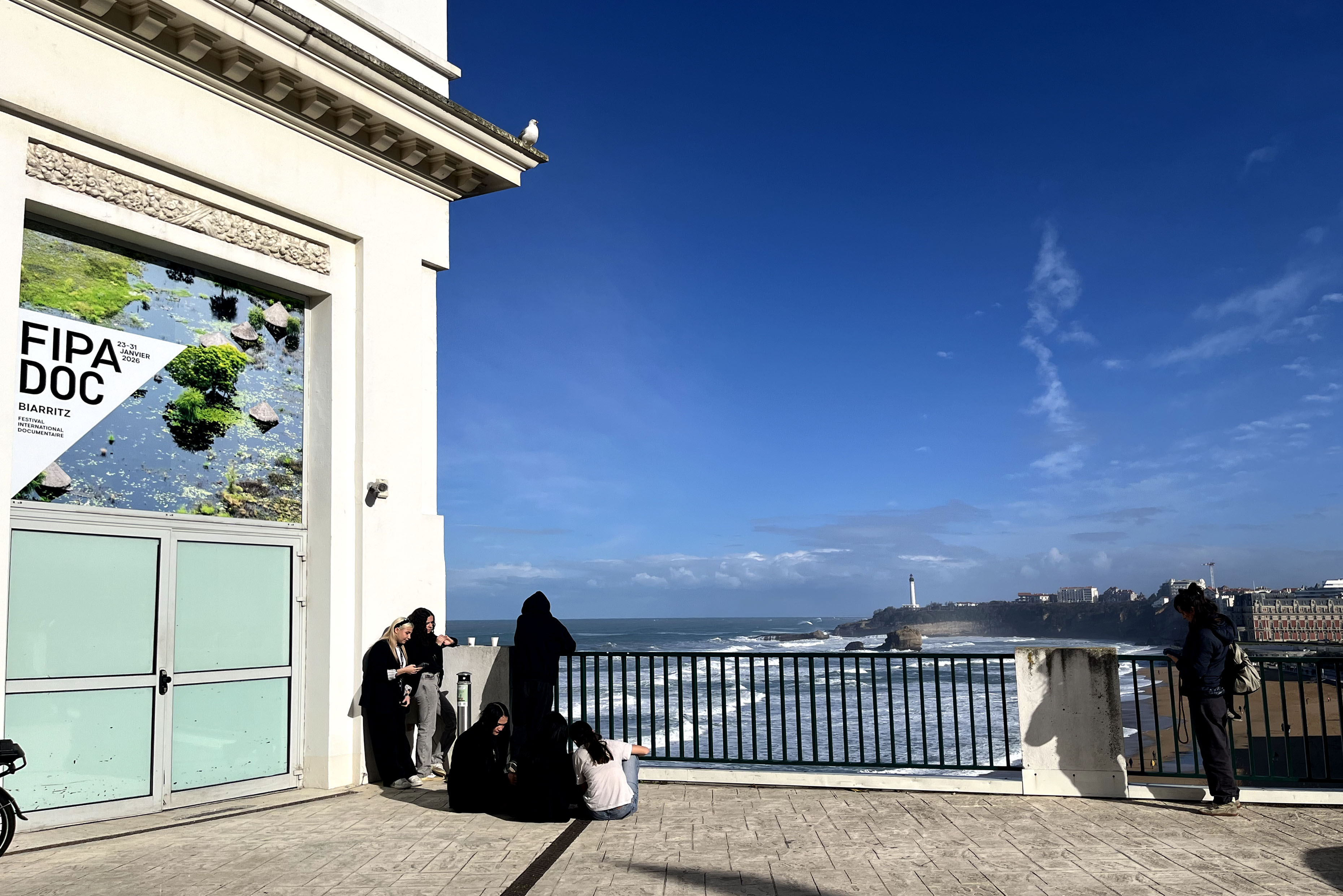 Overlooking crashing waves, a small group of people relax in front of a FIPADOC poster.