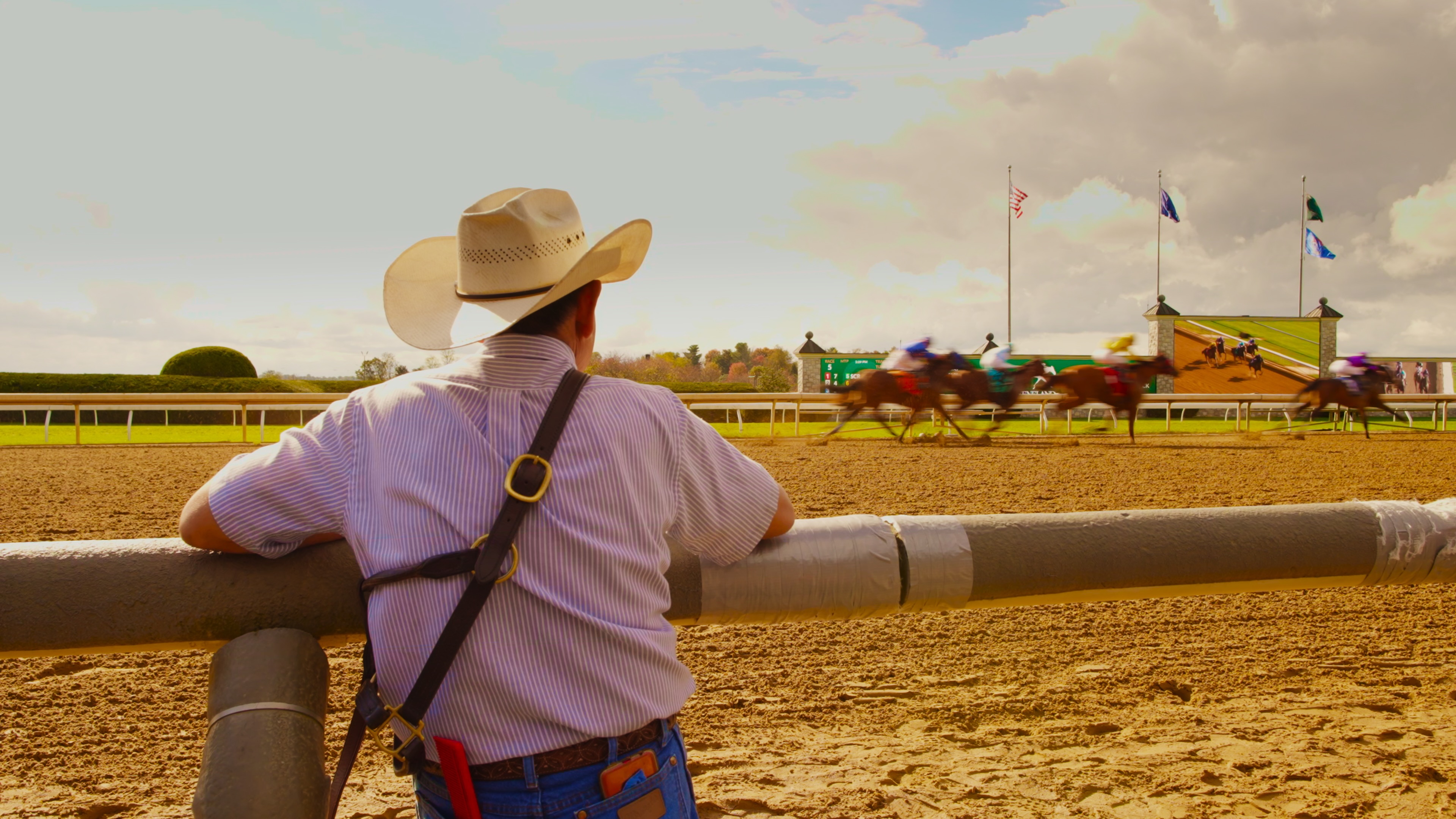 A man in a cowbow hat seen from behind is watching a horse race happening across from him